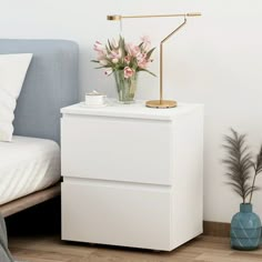 a white night stand next to a bed with flowers on it and a blue vase