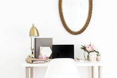 a white desk topped with a laptop computer next to a gold framed mirror and pink flowers