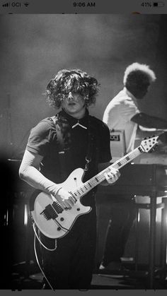 a man with curly hair playing an electric guitar in front of two other men on stage