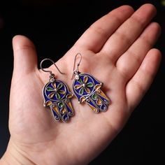 handmade Amazigh Hamsa earrings, crafted from sterling silver and adorned with vibrant enamel work. The Hamsa, a symbol of protection and good fortune, is intricately designed with traditional Moroccan Amazigh patterns. Product Details: Symbolism: The Hamsa symbol, known for its protective qualities, is a central motif in Amazigh culture, symbolizing blessings, power, and strength. Material: High-quality sterling silver and enamel. Dimensions: Width - 2.2 cm (0.87 inches) | Length - 5 cm (1.97 inches) Weight: 9.4 grams NOTE: Each pair of earrings is handcrafted, which may result in slight variations. The natural patina on the sterling silver can be left for an antique look or polished for a brighter shine. Please note that lighting can affect the appearance of the colors. Hamsa Symbol, Design Marocain, Hamsa Earrings, Good Fortune, Ethnic Jewelry, Earrings Sterling Silver, Sterling Earrings, Favorite Things Gift, Jewelry Earrings Dangle