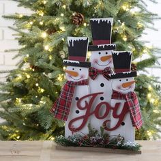 three snowmen standing in front of a christmas tree with the word joy written on it