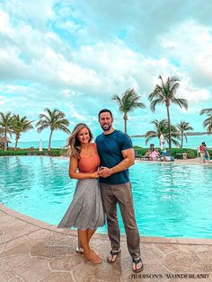 a man and woman standing in front of a swimming pool with palm trees behind them