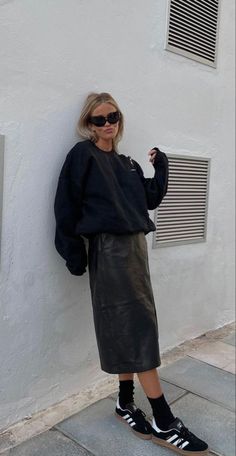 a woman leaning against a white wall with her hands on her hips and wearing black sneakers