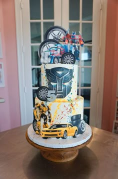 a three tiered cake decorated with cars and trucks is on a table in front of a window