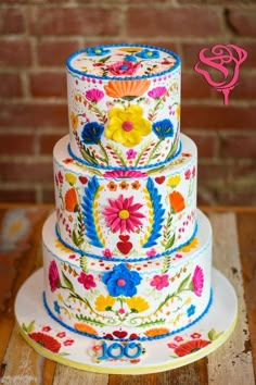 three tiered cake decorated with colorful flowers on a wooden table next to a brick wall