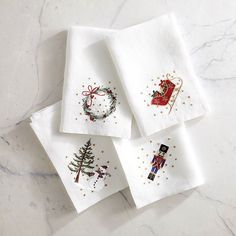 four white christmas napkins with embroidered designs on them, sitting on a marble surface