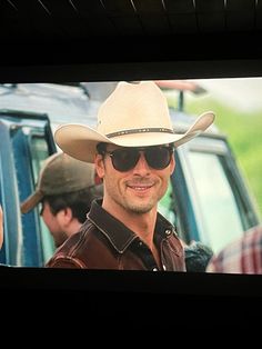 a man wearing a cowboy hat and sunglasses in front of a tv screen with people standing around