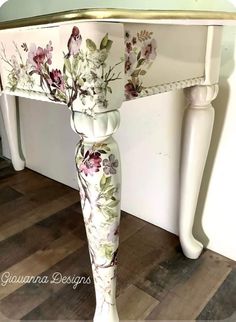 a white table with flowers painted on it and gold trim around the top, sitting on a hard wood floor