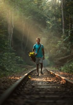 a man is walking down the railroad tracks with flowers in his hand and sunlight coming through the trees behind him