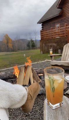 a drink sitting on top of a wooden table next to a fire pit in front of a log cabin