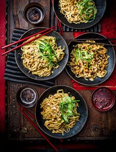 three plates of noodles with chopsticks on a wooden table next to bowls of sauce