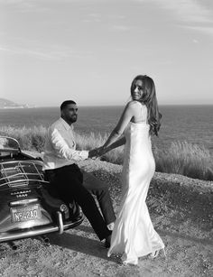 a man and woman holding hands while sitting on a motorcycle near the ocean, black and white photograph