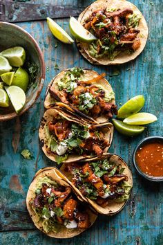 three tacos with meat, salsa and limes on a blue wooden table next to two bowls