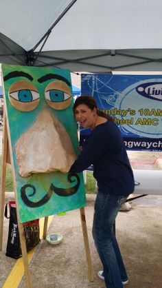 a man is standing next to a painting on an easel with a face painted on it