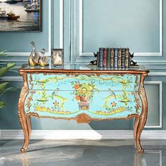 an ornate painted chest with books on top in front of a blue wall and painting