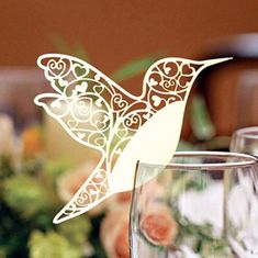 a paper bird sitting on top of a table next to two wine glasses with flowers in the background