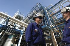 two men standing in front of an oil and gas plant with blue sky behind them