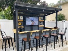 an outdoor bar with stools and a television on the wall above it, in front of a fence