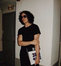 a man in black shirt and sunglasses standing next to white wall holding a book case