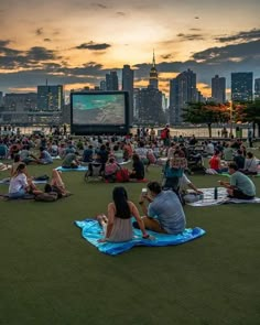 many people are sitting on the grass in front of an outdoor movie screen at sunset