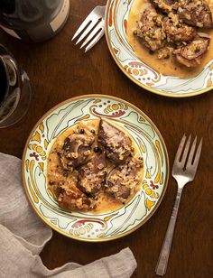 two plates with meat and gravy on them next to silverware, wine glasses and utensils