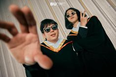 two people dressed in graduation gowns and sunglasses making the v sign with their hands