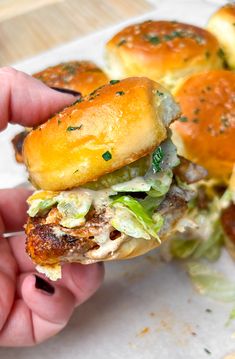 a hand is holding a sandwich with meat and lettuce on it, while other sliders are in the background