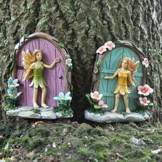 two fairy figurines are sitting on the ground next to a tree with flowers