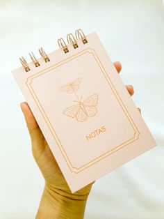 a hand holding a notepad with a butterfly on it and the words notas written in gold ink