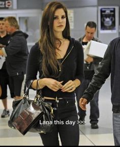 a woman with long hair walking through an airport carrying a black purse and looking at the camera