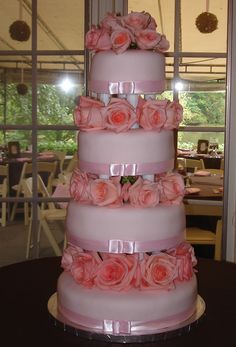 a three tiered wedding cake with pink roses on the top and bottom, in front of a window