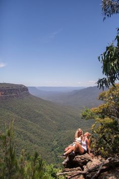 two people sitting on the edge of a cliff looking out over a valley with mountains in the background