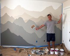 a man standing in front of a wall with mountains painted on it and paint buckets next to him