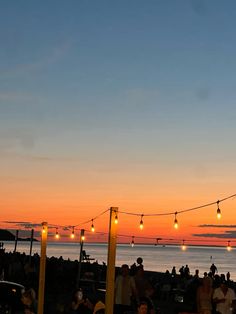 many people are sitting at tables near the water with lights strung over them and cars parked on the street