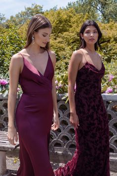 two women in long dresses standing next to each other on a stone bench with pink flowers behind them