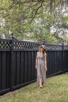 a woman standing in front of a black fence wearing a dress and hat with her hands on her hips