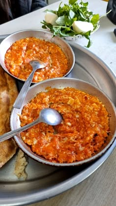 three bowls of food on a silver plate with spoons in each bowl and bread