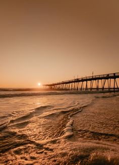 the sun is setting over an ocean with a long pier in the distance and waves crashing on the shore