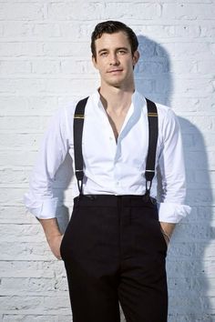 a man standing in front of a brick wall wearing suspenders and a white shirt