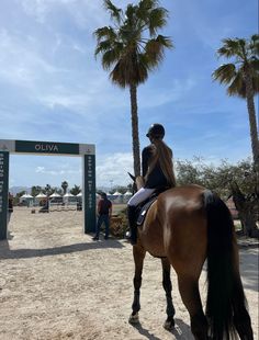 a woman riding on the back of a brown horse down a sandy beach covered in palm trees