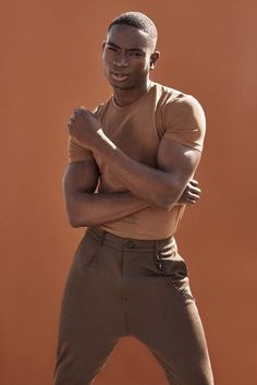 a man standing with his arms crossed in front of an orange background wearing brown pants and a tan t - shirt