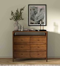 a wooden dresser sitting in front of a painting on the wall next to a potted plant