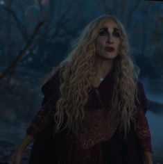 a woman with long blonde hair and makeup looks at the camera in a dark forest