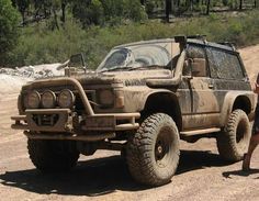 a man standing next to a dirty truck on top of a dirt road with trees in the background