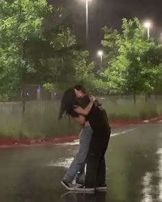 two people kissing on a skateboard in the rain at night with street lights behind them