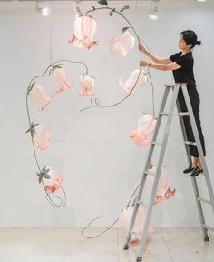 a woman standing on a ladder next to a wall with pink flowers hanging from it