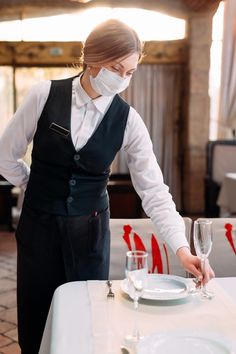 a woman wearing a face mask standing at a table with plates and glasses on it