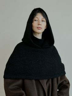 a woman wearing a black and brown cloak over her head, standing in front of a white wall