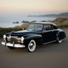 an old black car is parked on the side of the road near the water and hills