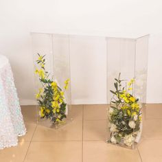 three clear vases filled with yellow flowers on top of a tiled floor next to a white table cloth